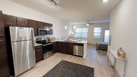 This single-floor space showcases a thoughtfully designed open-concept layout that seamlessly integrates the kitchen, living, and sleeping areas into one cohesive living environment. The kitchen is a standout feature, outfitted with rich dark espresso cabinetry, granite countertops, and a full suite of stainless steel appliances including a refrigerator, electric range, microwave, and dishwasher. A peninsula with pendant lighting serves as both a functional workspace and a natural divider between the kitchen and the rest of the living area. Track lighting above the kitchen provides focused illumination, while a ceiling fan in the main living area promotes comfortable airflow. Light-toned hardwood flooring runs throughout the space, lending a clean, contemporary aesthetic that ties the open floor plan together. Large windows along the far wall allow generous natural light to fill the interior, creating a bright and welcoming ambiance. A sleeping area is integrated into the open layout near the windows, benefiting from the same natural light and airy feel. The overall design balances modern finishes with a practical, efficient use of space, making it a functional and visually appealing living environment.