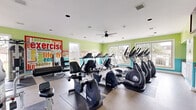This fitness center in Montgomery Village, Maryland offers a thoughtfully designed workout space spread across a single open floor. The room is immediately eye-catching, with bold lime green and teal walls accented by large, colorful word-art murals centered around themes of exercise and healthy living, creating an energizing and motivating atmosphere.

The cardio zone is well-stocked, featuring a row of Spirit treadmills, multiple elliptical trainers, and stationary recumbent bikes, all arranged to maximize floor space while allowing comfortable movement between machines. Rubber floor mats beneath each piece of equipment provide safety and stability. The strength training area includes a multi-station cable machine, a weight bench, and additional resistance equipment, offering a comprehensive selection for full-body workouts.

Large windows along the walls flood the space with natural light and provide views of the surrounding outdoor area. Ceiling fans assist with air circulation, keeping the environment comfortable during intense sessions. Recessed lighting in the white ceiling supplements natural light for consistent illumination throughout the day.

The flooring is a clean, durable tile that transitions seamlessly across the space. A wall-mounted television is also present for entertainment during workouts. Overall, the fitness center delivers a vibrant, functional, and well-maintained environment suited for a wide range of exercise routines.