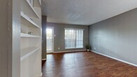 This single-story residence offers a well-designed layout with consistent hardwood-style flooring and a cohesive neutral gray color palette throughout. The living area is open and inviting, featuring large windows with vertical blinds that allow abundant natural light to fill the space. A built-in shelving unit adds functional storage and architectural character near the entryway.

The kitchen is a standout feature, outfitted with rich dark wood cabinetry, granite-style countertops, a tile mosaic backsplash, and black appliances including a refrigerator, electric range, and dishwasher. Ample upper and lower cabinetry provides generous storage, and the galley-style layout promotes efficient use of the space.

The bedrooms are well-proportioned, each finished with the same warm hardwood-style flooring and neutral wall tones. One bedroom includes a ceiling fan with light fixture, while windows with blinds offer natural light and privacy. The bathroom is thoughtfully arranged, featuring a vanity with a countertop sink, dark cabinetry, a tiled shower-tub combination, and a walk-in closet for additional storage. Textured ceilings are present throughout the home, adding a classic architectural touch. Overall, the space presents a clean, functional, and comfortable living environment.