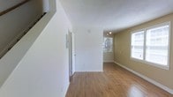 This two-story home presents a welcoming interior defined by clean lines and a straightforward, functional design. The main level features warm hardwood flooring that extends throughout the open living areas, complementing the crisp white walls and neutral-toned accent walls. A staircase is visible near the entry, providing access to the upper floor and adding vertical interest to the space.

The living area is generously proportioned, with multiple windows allowing abundant natural light to fill the room. The windows offer views of surrounding greenery, contributing to a bright and airy atmosphere. A track lighting fixture is mounted on the ceiling in the adjoining area, providing targeted illumination and a subtle modern touch.

White baseboards frame the flooring throughout, adding a clean, finished look to each room. Doorways lead to additional spaces off the main living area, suggesting a practical layout with good flow between rooms. The overall ambiance is neutral and move-in ready, offering a blank canvas that accommodates a variety of interior styles and furnishing choices across both levels of the home.