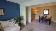 This single-story space showcases a thoughtfully designed open floor plan that connects the living area, dining space, and kitchen in a cohesive layout. The living area is anchored by a striking navy blue accent wall, which provides a bold contrast against the soft, neutral beige tones found throughout the rest of the space. Plush wall-to-wall carpeting in a warm tan hue extends across the main living and dining areas, adding comfort underfoot and contributing to the space's cozy ambiance. A decorative potted plant near the seating area introduces a natural element that softens the overall aesthetic. The dining area accommodates a table with seating and is positioned adjacent to the kitchen, which is partially visible through an open passthrough or entry. The kitchen features warm wood-toned cabinetry and is illuminated by warm ambient lighting, lending it an inviting atmosphere. The layout flows naturally between areas, making the space feel connected and functional. Neutral wall colors in the kitchen and dining areas complement the bolder design choices in the living space, creating a balanced and harmonious interior throughout.