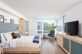 This contemporary single-floor studio is thoughtfully designed to maximize both function and comfort. Light hardwood flooring flows throughout the entire unit, creating a cohesive and airy feel. An entry hallway, framed by a sleek sliding barn door, leads into the open living space and features a dedicated closet area with a curtained enclosure.

The kitchen is outfitted with dark wood-grain cabinetry extending to the ceiling, a stainless steel induction cooktop, a double undermount sink, a stainless steel dishwasher, and a full-size refrigerator — providing a complete and efficient cooking environment. Ample counter space and upper cabinetry ensure generous storage throughout.

The combined living and sleeping area is bright and open, anchored by a full bed set against a fluted blue accent wall with integrated wall sconces. Large windows with horizontal blinds allow natural light to fill the room while offering views of the surrounding neighborhood. A seating area with a modern lounge chair and a compact dining table positioned near the windows complete the living space.

The bathroom features dark-toned vanity cabinetry with a wide countertop, a large mirror with overhead lighting, hardwood-style flooring, and a shower enclosed by a curtain. The overall design aesthetic is clean, minimal, and modern throughout.