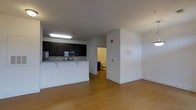 This single-floor space is designed with an open-concept layout that integrates the kitchen and living area into one cohesive environment. The kitchen is outfitted with dark espresso cabinetry that contrasts cleanly against the white walls, offering ample storage. Stainless steel appliances, including a microwave, range, and refrigerator, provide modern functionality. A peninsula-style breakfast bar with light granite countertops extends into the living space, creating a natural transition between cooking and lounging areas and offering casual seating.

The living area is generously proportioned, featuring warm-toned hardwood flooring that runs throughout and contributes to an inviting atmosphere. White walls and a neutral color palette keep the space feeling bright and airy. Overhead fluorescent lighting provides even illumination throughout the main living area.

A doorway visible from the main space leads to an adjacent room, likely a bathroom, suggesting additional utility within the floor plan. The overall design is clean and contemporary, emphasizing practicality and openness, making the space well-suited for everyday living.