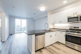 This contemporary single-floor unit offers a clean, modern aesthetic with thoughtfully designed spaces and quality finishes throughout. The open-concept layout seamlessly connects the kitchen and living area, creating a bright and functional living environment. Light wood-tone luxury vinyl plank flooring runs throughout the main areas, contributing to a cohesive and welcoming atmosphere.

The kitchen is a standout feature, centered around a peninsula with dark countertops that provide generous prep and dining space. White flat-panel cabinetry with matte black hardware extends to the ceiling, maximizing storage. Stainless steel appliances — including a gas range, over-the-range microwave, refrigerator with ice maker, and dishwasher — complete the well-appointed culinary space. Under-cabinet lighting adds a polished finishing touch. The adjacent living area benefits from a sliding glass door that invites natural light and provides access to an outdoor balcony.

The bedroom is a comfortable retreat featuring a large window with blinds and direct access to a walk-in closet with built-in shelving and hanging rods. The bathroom is finished in a clean, neutral palette, featuring a soaking tub with tiled surround, a toilet, and a wide vanity with dark countertops, white cabinetry, and a large wall mirror with bright overhead lighting.