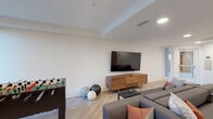 Located at 12041 Wilshire Blvd in Los Angeles, this single-floor amenity space offers a well-appointed communal lounge designed for both relaxation and socializing. The interior features a clean, contemporary aesthetic throughout, with light wood-look flooring, white walls, and recessed lighting that create a bright, welcoming atmosphere.

The main lounge area is anchored by a large wall-mounted flat-screen television set above a warm walnut media console, paired with a generously sized sectional sofa accented with coral and patterned throw pillows. A foosball table and fitness balls nearby add a recreational element, making the space versatile for leisure and entertainment.

A dedicated dining nook offers a striking visual contrast, featuring a vibrant, large-scale geometric mural in bold tones of pink, purple, gold, and blue. A square table with modern black chairs sits beneath recessed lighting, with glass doors opening to an outdoor area with visible greenery.

The kitchenette is finished with textured gray cabinetry, white quartz countertops, a stainless steel double sink, under-cabinet lighting, and a built-in microwave — providing practical functionality to complement the social spaces. Together, these areas form a cohesive, stylish communal environment well-suited for an urban Los Angeles setting.