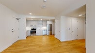 This single-floor unit presents a cohesive, modern aesthetic defined by crisp white walls and warm light wood flooring that runs continuously throughout the space. The open-concept kitchen is a standout feature, equipped with flat-front gray cabinetry extending floor to ceiling, stainless steel appliances including a range, dishwasher, and refrigerator, complemented by a white subway tile backsplash and light-toned countertops. Recessed lighting illuminates the kitchen area effectively. The living area adjoins the kitchen, offering an open and versatile layout with clean sightlines. Additional rooms provide flexible use, with at least one bedroom benefiting from a large window overlooking a lush, green outdoor setting, allowing natural light and garden views. The bathroom features a navy-toned vanity with ample storage, a white countertop, and a large mirror. A dedicated in-unit laundry area with a stacked washer and dryer is conveniently located nearby. Throughout the unit, white baseboards, minimalist hardware, and a neutral palette contribute to a clean, contemporary atmosphere.