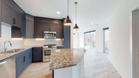 This contemporary single-floor residence showcases a thoughtfully designed open-concept layout that seamlessly connects the kitchen and living areas. The kitchen serves as the centerpiece, featuring dark charcoal cabinetry that extends to the ceiling, a textured white tile backsplash, and a generously sized island topped with striking granite countertops. Stainless steel appliances, including a gas range, built-in microwave, and dishwasher, complement the space, while two pendant lights with matte black fixtures provide focused task lighting overhead. Light wood-style flooring runs throughout the main living areas, enhancing the bright, airy atmosphere created by large floor-to-ceiling windows overlooking urban skyline views.

The bedrooms are finished with plush carpeting and crisp white walls, with several rooms offering large windows or sliding glass doors that open to a private balcony with city views. The primary bathroom features a double-sink granite vanity with dark cabinetry, a glass-enclosed walk-in shower with tile surround, and light stone flooring. A secondary bathroom includes a soaking tub with tiled surround and matching granite vanity finishes, maintaining a cohesive aesthetic throughout the residence.