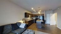 This single-floor space showcases an open-concept layout that combines the living and kitchen areas into one cohesive, functional environment. The kitchen is well-appointed with rich dark wood cabinetry, granite-style countertops, and black appliances including a range, microwave, and refrigerator. A peninsula island with seating provides an ideal spot for casual dining or entertaining, illuminated by stylish pendant lighting overhead. Recessed ceiling lights ensure the space is well-lit throughout. The living area flows naturally from the kitchen, featuring warm wood-look flooring that ties the two spaces together. Neutral-toned walls and a clean white ceiling contribute to a bright and welcoming ambiance. The living room offers comfortable seating and is accented by a classic table lamp, adding a warm, homey touch. The overall design balances practicality with a modern aesthetic, making it a versatile and inviting space. A hallway visible in the background suggests additional rooms beyond the main open area, rounding out this thoughtfully designed single-level layout.