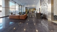 Located on Wilshire Boulevard in the heart of Los Angeles, this ground-floor lobby makes an immediate impression with its sophisticated, contemporary design. The space is defined by richly polished dark stone flooring that reflects the ambient and natural light, creating a luminous, expansive feel throughout. A striking transition from the entry corridor to the main lobby area showcases a combination of dark and light materials — including large-format marble wall cladding with warm gold trim accents — lending the space an air of refined elegance. The entry corridor features elevator banks framed by veined marble panels, complemented by recessed and pendant lighting that guides visitors seamlessly into the main reception area. The open lobby is flooded with natural light through floor-to-ceiling glass walls, offering views to the street beyond. A sculptural geometric accent wall in white adds texture and visual interest, while a sleek reception desk anchors the space. Comfortable contemporary seating, including a warm-toned sectional sofa, invites visitors to relax. Tasteful greenery and modern light fixtures complete the polished, welcoming ambiance of this well-appointed Los Angeles lobby.