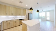 This well-appointed one-bedroom, one-bathroom apartment in Roanoke, TX offers a thoughtfully designed layout with modern finishes throughout. The open-concept kitchen and living area serves as the heart of the home, featuring light wood cabinetry, white quartz countertops, a full suite of stainless steel appliances, and a central island with pendant lighting overhead. Light hardwood-style flooring extends through the main living areas, enhancing the bright, airy atmosphere. The living room is bathed in natural light from large windows and a glass door that opens directly onto a private balcony, offering outdoor views of the surrounding landscape. A ceiling fan adds comfort to the space. The bedroom is carpeted for warmth, equipped with a ceiling fan, and benefits from a large window with outdoor views. The full bathroom features a soaking tub with tiled surround, wood-toned vanity with quartz countertop, and direct access to a walk-in closet with built-in shelving. Additional built-in open shelving is visible near the kitchen area, providing practical storage. An in-unit laundry area completes the functional layout of this clean, contemporary residence.