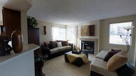 This single-floor residence offers a comfortable and thoughtfully arranged living space designed for both relaxation and everyday functionality. The living room serves as the heart of the home, anchored by a tiled fireplace that adds warmth and a classic focal point to the room. A wall-mounted display piece above the fireplace complements the space's curated aesthetic. Large windows along one wall allow generous natural light to filter in, brightening the neutral-toned interior and creating an airy, open feel. The living area is finished with plush carpeting that adds texture and comfort underfoot, while tile flooring near the fireplace provides a clean visual transition. The room accommodates ample seating, centered around a dark upholstered sofa accented with earth-toned throw pillows, and a coordinating ottoman that doubles as a coffee table. Lush indoor plants are thoughtfully placed throughout, adding a natural, lively element to the space. The textured ceiling adds subtle architectural character. Overall, the space presents a warm and inviting ambiance with a cohesive color palette of neutral tones, deep browns, and soft greens that lend a grounded, homey feel throughout.
