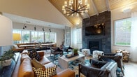 Located at 115 Dean Ave in Franklin, Massachusetts, this single-story community clubhouse showcases a thoughtfully designed interior that blends contemporary and rustic elements throughout its open, flowing layout. The architecture is immediately defined by dramatically vaulted ceilings clad in warm wood planking, supported by exposed beams and illuminated by striking statement chandeliers that anchor each zone of the space.

The main lounge area centers around a floor-to-ceiling dark stone fireplace with a mounted flat-screen television above, surrounded by leather seating, patterned ottomans, and a vibrant blue area rug. Large windows throughout flood the space with natural light and offer views of the surrounding greenery. A decorative laser-cut wood screen panel and sliding barn door add architectural interest while subtly dividing areas.

The kitchen features bold teal cabinetry, a white quartz island with bar seating, industrial pendant lighting, and stainless steel appliances, including a coffee station. Adjacent dining and lounge areas accommodate both casual gatherings and collaborative work, complete with a long communal table and built-in bench seating.

A dedicated game room offers a full-size billiards table, arcade machines, and geometric pendant lighting beneath its own vaulted ceiling, providing a lively entertainment hub. Light wood-tone flooring ties the entire space together cohesively.