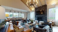 Located at 115 Dean Ave in Franklin, Massachusetts, this single-story community clubhouse showcases a thoughtfully designed interior that blends contemporary and rustic elements throughout its open, flowing layout. The architecture is immediately defined by dramatically vaulted ceilings clad in warm wood planking, supported by exposed beams and illuminated by striking statement chandeliers that anchor each zone of the space.

The main lounge area centers around a floor-to-ceiling dark stone fireplace with a mounted flat-screen television above, surrounded by leather seating, patterned ottomans, and a vibrant blue area rug. Large windows throughout flood the space with natural light and offer views of the surrounding greenery. A decorative laser-cut wood screen panel and sliding barn door add architectural interest while subtly dividing areas.

The kitchen features bold teal cabinetry, a white quartz island with bar seating, industrial pendant lighting, and stainless steel appliances, including a coffee station. Adjacent dining and lounge areas accommodate both casual gatherings and collaborative work, complete with a long communal table and built-in bench seating.

A dedicated game room offers a full-size billiards table, arcade machines, and geometric pendant lighting beneath its own vaulted ceiling, providing a lively entertainment hub. Light wood-tone flooring ties the entire space together cohesively.