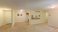 This single-floor space features an open-concept layout that seamlessly connects the living area to the kitchen. The living area is finished with wall-to-wall beige carpet and neutral cream-colored walls, creating a clean and understated aesthetic. Recessed structural framing defines the boundary between the living space and the kitchen without fully closing off the two areas, maintaining an open and airy feel throughout.

The kitchen is accessible via a pass-through counter that opens directly into the living area, providing both visual connectivity and functional convenience. The kitchen is equipped with a white gas range and a white refrigerator, complemented by dark wood cabinetry mounted above. A single-basin sink is positioned along the pass-through wall, and the kitchen surfaces feature a neutral countertop finish. The overhead lighting in the kitchen provides direct illumination to the workspace.

Flush-mount ceiling lighting serves the main living area, and white baseboards run consistently along the perimeter of the space. The overall design is practical and cohesive, with a neutral palette that offers a versatile foundation for a variety of interior arrangements.