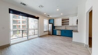 This single-floor residence showcases a contemporary design highlighted by a striking color palette and quality finishes throughout. The open-concept living and kitchen area is anchored by light wood-tone flooring and recessed lighting, creating a bright, airy atmosphere. Large sliding glass doors lead to a private balcony, flooding the space with natural light and offering views of the surrounding building exterior.

The kitchen is a standout feature, boasting bold teal lower cabinetry paired with crisp white upper cabinets, gold-toned hardware, and granite countertops. A light gray subway tile backsplash complements the stainless steel appliances, which include a refrigerator, electric range with microwave, and dishwasher. Open wood shelving adds a modern, functional touch.

The bedrooms are carpeted for comfort and equipped with ceiling fans. The primary bedroom connects directly to a walk-in closet and an en-suite bathroom. The bathroom continues the teal cabinetry theme, featuring a granite vanity top, a backlit vanity mirror, and a full tub-shower combination with clean white tile surround and brushed nickel fixtures throughout.