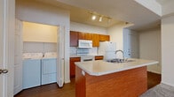 Located at 1350 W Van Buren in Phoenix, Arizona, this single-floor unit offers a well-organized, functional layout designed for comfortable everyday living. The kitchen is a standout feature, outfitted with warm-toned wood cabinetry, white appliances including an electric range, over-the-range microwave, and refrigerator, as well as a peninsula island with a double stainless steel sink and light countertops. Track lighting illuminates the kitchen area, while durable hardwood-style flooring runs throughout. A dedicated in-unit laundry area with a full-size washer and dryer is conveniently accessible directly from the kitchen. The open living area flows naturally from the kitchen and is finished with plush carpeting, recessed lighting, and a ceiling fan. Vertical blinds line the sliding door, allowing for adjustable natural light. The bedroom offers generous proportions, soft carpet flooring, and multiple windows dressed with vertical blinds that bring in ample natural light while maintaining privacy. The bathroom features a full tub-shower combo with white tile surround, complemented by wood-style flooring. Clean white walls throughout the unit create a bright, neutral backdrop, making the space feel open and easy to personalize.