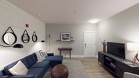 This single-floor residence offers a thoughtfully designed layout that balances comfort and modern style throughout. The living area serves as the heart of the space, highlighted by a striking white panel accent wall adorned with decorative round mirrors and swing-arm wall sconces. Wood-look flooring runs throughout the main areas, complementing the neutral gray and white color palette that gives the space a clean, contemporary feel.

The kitchen is well-equipped and highly functional, featuring a two-tone design with crisp white upper cabinetry and dark lower cabinets, a classic subway tile backsplash, and stainless steel appliances including a refrigerator, range, and microwave. A granite-topped island with an undermount sink doubles as a breakfast bar, with pendant lighting above and a window that allows natural light to flow in.

The bedroom is a serene retreat with plush carpeting, neutral walls, and a generous walk-in closet. A decorative floating shelf above the headboard area adds character, while well-placed lighting creates a warm, inviting ambiance.

The bathroom connects conveniently to the bedroom and features wood-look flooring, dark cabinetry with a granite vanity top, a round mirror, and pendant lighting, offering a polished and practical finish to this well-rounded residence.