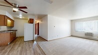 This single-floor unit offers a practical and functional layout across two bedrooms, one bathroom, and a combined kitchen and living area. The open living room is generously sized, featuring neutral carpet flooring, a ceiling fan, and a large window dressed with vertical blinds that allows natural light to fill the space. A baseboard heating system and a wall-mounted air conditioning unit provide year-round climate control. The living area transitions seamlessly into the kitchen, which features warm wood cabinetry, granite-style laminate countertops, a double sink, and white appliances including a refrigerator and range. Hard surface wood-look flooring runs throughout the kitchen, complementing the cabinetry tones. The primary bedroom is a comfortable size, offering a walk-in closet with built-in shelving and a window with vertical blinds. The second bedroom includes a sliding wood closet door with a shelf and hanging rod. The bathroom features a wide vanity with a granite-style countertop, an oval sink, and warm wood cabinetry. Throughout the unit, wood-trimmed doors and warm-toned finishes create a cohesive, understated aesthetic.