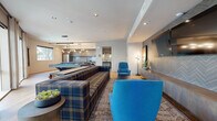 Located at 222 W Boston St in Chandler, Arizona, this thoughtfully designed single-floor amenity space offers a well-rounded social environment across three distinct zones. The main lounge area serves as the centerpiece, furnished with a bold plaid sectional sofa and vibrant blue accent chairs atop a layered area rug. A textured herringbone-patterned feature wall anchors a wall-mounted flat-screen TV, complemented by a sleek floating media console, contemporary floor lamp, and curated décor. Light wood flooring runs throughout, creating a warm and cohesive aesthetic. Recessed lighting provides even illumination across the entire space. The kitchen area is outfitted with rich wood cabinetry, stainless steel appliances including a full-size refrigerator, a striking stone-veined island with bar seating, and a teal glass tile backsplash that adds a vibrant pop of color. An adjacent restroom provides added convenience. The game room features a modern pool table as its focal point, bathed in natural light from large floor-to-ceiling windows and additional wall windows that frame views of palm trees and the surrounding outdoor amenities. A wall-mounted shuffleboard game adds to the recreational appeal. Together, these zones create a versatile, inviting gathering space well-suited for both relaxation and entertainment.
