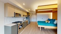 Located in Portland's vibrant Woodstock neighborhood, this modern studio unit offers a well-conceived open-plan layout that maximizes every square foot through smart, contemporary design. Light wood-look flooring runs throughout, lending warmth and continuity across the entire space.

The kitchen is a standout feature, outfitted with stainless steel appliances including a gas range, over-the-range microwave, dishwasher, and refrigerator. Two-tone cabinetry pairs light wood upper cabinets with sage-green lower drawers, complemented by a geometric white tile backsplash and a sleek farmhouse-style stainless steel sink with a matte black faucet — a refined combination of modern and functional design.

The sleeping area is anchored by a custom built-in bed platform framed by a striking warm wood panel accent wall and an architectural wood-slat ceiling canopy with integrated ambient lighting, adding depth and a boutique-hotel quality to the space. A large window bathes the living area in natural light and offers views of the surrounding tree-lined neighborhood.

The bathroom is clean and minimal, featuring a wood-toned vanity with white countertop, a soaking tub with shower, and matte black fixtures throughout. A stacked washer and dryer unit is conveniently integrated directly off the bathroom, providing in-unit laundry without sacrificing usable space.