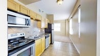 This single-floor space showcases a functional, open layout where the kitchen flows seamlessly into the living area. The kitchen is well-equipped with a full suite of appliances, including a stainless steel refrigerator, electric range, built-in microwave, and dishwasher. Light wood cabinetry lines the walls, providing generous storage, while a decorative tile backsplash adds texture and visual interest behind the cooking and prep areas. Dark countertops contrast neatly with the cabinetry, and a double-basin sink sits beneath under-cabinet lighting for practical use. The kitchen floor is finished in neutral ceramic tile, which transitions into carpeted flooring in the adjoining living area. The living space is bright and airy, benefiting from multiple windows dressed with horizontal blinds that allow natural light to filter throughout. Warm beige wall tones carry through both areas, contributing to a cohesive and inviting atmosphere. Flush-mount ceiling lighting provides additional illumination, ensuring the space remains well-lit. Overall, the layout offers a practical and comfortable living environment with a clean, neutral aesthetic throughout.