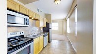 This single-floor space showcases a functional, open layout where the kitchen flows seamlessly into the living area. The kitchen is well-equipped with a full suite of appliances, including a stainless steel refrigerator, electric range, built-in microwave, and dishwasher. Light wood cabinetry lines the walls, providing generous storage, while a decorative tile backsplash adds texture and visual interest behind the cooking and prep areas. Dark countertops contrast neatly with the cabinetry, and a double-basin sink sits beneath under-cabinet lighting for practical use. The kitchen floor is finished in neutral ceramic tile, which transitions into carpeted flooring in the adjoining living area. The living space is bright and airy, benefiting from multiple windows dressed with horizontal blinds that allow natural light to filter throughout. Warm beige wall tones carry through both areas, contributing to a cohesive and inviting atmosphere. Flush-mount ceiling lighting provides additional illumination, ensuring the space remains well-lit. Overall, the layout offers a practical and comfortable living environment with a clean, neutral aesthetic throughout.