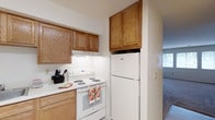Village East Apartments presents a well-appointed single-floor residence designed for comfortable everyday living. The kitchen is a practical and inviting space, featuring warm honey-toned wood cabinetry that provides generous storage both above and below the countertops. White appliances, including a full-size refrigerator and an electric range with coil burners, are neatly integrated into the layout. The light-colored countertops and under-cabinet lighting create a clean, functional cooking environment. The kitchen flows naturally into the adjacent living area, which benefits from natural light streaming in through a window, creating a bright and welcoming atmosphere. The single-floor layout ensures easy accessibility and a seamless transition between living spaces. The neutral color palette throughout the residence lends itself to a versatile interior style, making it easy to personalize. Overall, this residence at Village East Apartments combines practical design with a cozy ambiance, offering a comfortable and functional home environment.