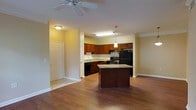 This single-floor space features an open-concept layout that seamlessly connects the living area to the kitchen, creating a spacious and functional environment. The kitchen is a standout feature, showcasing rich dark-cherry cabinetry that extends to the upper walls, providing generous storage throughout. Granite countertops add an elegant touch, complemented by a matching center island that serves as both a prep surface and a natural gathering point. The kitchen is fully equipped with black appliances, including a refrigerator, range, and dishwasher, and is illuminated by stylish pendant lighting that hangs above the island alongside a bright overhead fixture. Crown molding lines the ceiling throughout, adding a refined architectural detail to the space. The living area is finished with warm hardwood flooring that flows continuously from room to room, enhancing the cohesive feel of the open layout. A ceiling fan in the living area provides both comfort and light. Neutral warm-toned walls contribute to an inviting ambiance, while an entry door is conveniently accessible from the main living space. Overall, the design balances practicality with tasteful finishes, making it a comfortable and well-detailed single-level home.