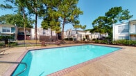 Located in Lafayette, Louisiana, this community amenity space offers residents a well-rounded set of recreational facilities designed to support an active lifestyle. The centerpiece of the outdoor area is a generously sized swimming pool with clear blue water, surrounded by a brick-edged deck ideal for lounging. The pool area is enclosed by a black iron fence and framed by tall, mature pine trees that provide natural shade and a lush backdrop against the Louisiana sky.

Adjacent to the pool, the fitness center is accessible via a direct exterior door, making transitions between indoor and outdoor workouts seamless. Large windows line one wall of the fitness center, allowing abundant natural light and direct views of the pool area, creating an open and energizing workout environment. The interior features patterned carpet flooring and a textured ceiling with recessed fluorescent lighting.

The fitness center is equipped with a variety of cardio machines, including ellipticals, treadmills, and a stationary bike, as well as a multi-station cable weight machine positioned against a teal accent wall. The space is well-organized, providing ample room to move between equipment comfortably. Together, the pool and fitness center form a cohesive, functional amenity hub that enhances the overall living experience at this Lafayette community.