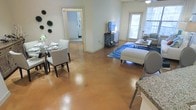 This single-story space is defined by its striking polished concrete floors, which carry a warm amber tone throughout the open-concept main living area, creating a cohesive and visually distinctive foundation. The layout seamlessly integrates a dining area and a living space, promoting an airy, connected feel. The dining area accommodates a glass-top table with white upholstered chairs, while decorative greenery accents the adjacent wall. The living area features a seating arrangement centered around a blue area rug, adding a pop of color against the warm flooring. A media console and television anchor one wall, while natural light filters in through windows dressed with blinds, brightening the space. A ceiling fan overhead provides both function and subtle style. Granite countertops are partially visible at the edge of the frame, hinting at a nearby kitchen with quality finishes. An open doorway leads to an additional room, suggesting a flexible and practical floor plan. The soft, neutral wall tones complement the warm flooring, creating an overall ambiance that is bright, open, and welcoming.
