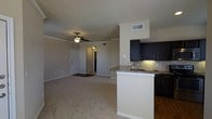 This single-story residence showcases an open-concept design that seamlessly connects the living area and kitchen into one cohesive space. The living area is finished with neutral-toned carpet, contributing to a warm and inviting atmosphere. A ceiling fan with a light fixture is centrally positioned, providing both air circulation and ambient lighting. Crown molding accents the upper walls, adding a subtle architectural detail throughout.

The kitchen is defined by dark cabinetry that contrasts sharply against light-colored walls, creating a visually striking aesthetic. A peninsula with a granite-style countertop extends into the living space, offering additional workspace and a natural division between the two areas. The kitchen features a tile backsplash, under-cabinet lighting, and a sink integrated into the peninsula. Dark hardwood flooring grounds the kitchen area, distinguishing it from the carpeted living space.

A doorway visible from the living area leads further into the residence, while the front entry door is positioned along the adjacent wall. Neutral wall tones throughout the space enhance its bright, clean appearance and provide a versatile backdrop for furnishing.
