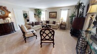 This single-story residence offers a thoughtfully designed living space that blends comfort and classic elegance. The living room serves as the centerpiece of the home, showcasing a generous, open layout finished with light neutral-toned walls and plush carpeted flooring that adds warmth underfoot. Large windows dressed with white curtains allow natural light to fill the room, creating a bright and inviting atmosphere throughout the day. The space is furnished with a coordinated arrangement of cream-upholstered sofas accented with rich, dark decorative pillows, complemented by dark mahogany-toned accent chairs featuring an intricate lattice back design. Classic end tables with warm-toned lamps add a refined ambiance, while a framed landscape painting serves as a tasteful focal point on the wall. Lush indoor plants, including a striking tropical variety, introduce a touch of natural greenery that enlivens the neutral palette. Built-in closet space and additional cabinetry contribute to the room's functional design. The overall ambiance is one of timeless, traditional elegance — a comfortable and well-composed space suited for both relaxed living and gracious entertaining.