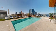 Perched atop a building in downtown San Diego's vibrant Little Italy neighborhood, this rooftop amenity deck offers a resort-style retreat with sweeping, unobstructed views of the San Diego skyline and beyond. The centerpiece of the space is a generously sized rectangular swimming pool with crystal-clear turquoise water, complemented by a separate spa — both finished with elegant tile accents and surrounded by smooth, light-colored concrete decking. An outdoor flat-screen television is mounted poolside, adding a modern entertainment element to the alfresco setting. Lush potted tropical plants and greenery are thoughtfully placed throughout the deck, softening the urban environment and enhancing the resort ambiance. Ample seating is available in the form of cushioned chaise lounges arranged along turf-lined sections of the deck, alongside patio furniture and market umbrellas for shade. Glass and metal railings line the perimeter, preserving the breathtaking panoramic views of the city's high-rises, waterfront, and open sky. The space enjoys abundant natural sunlight year-round, making it an ideal outdoor destination for relaxation in the heart of San Diego.