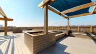Located at 585 North Ave in New Rochelle, New York, this rooftop deck offers a well-appointed outdoor space designed for both comfort and functionality. The deck is finished with durable composite decking in a warm gray tone, providing a clean and low-maintenance surface throughout. A prominent wood-framed pergola anchors the central gathering area, outfitted with teal shade sails that provide partial cover while allowing abundant natural light to filter through beneath open skies.

Built-in raised planters constructed from matching composite material are integrated into the layout, creating opportunities for greenery and garden use directly on the rooftop. Tiered seating areas accompany the planters, offering casual, built-in bench seating for occupants. The perimeter is defined by lattice-panel wood railings that provide safety while maintaining an open, airy feel with unobstructed views of the surrounding New Rochelle skyline and landscape.

A walkway runs along the building's exterior facade, finished in the same composite decking and accessible via large floor-to-ceiling glass sliding doors, seamlessly connecting the interior to the outdoor space. The modern tiled building exterior complements the natural wood tones of the pergola and railing structures. The overall design balances urban practicality with an inviting outdoor atmosphere, making this rooftop deck a distinctive and functional amenity.