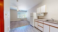 This single-floor unit offers an efficient and functional layout, combining the kitchen and living area into one open space. The kitchen is well-equipped with white cabinetry featuring wood-tone trim accents, a full-size refrigerator, electric range, built-in microwave, and a double-basin sink set into a continuous countertop. Ample upper and lower cabinet storage keeps the kitchen organized and practical. Light wood-style flooring runs through the kitchen area, transitioning into plush carpeting in the living space. The living area is brightened by a large window fitted with horizontal blinds, allowing generous natural light to fill the room while offering outdoor views. A ceiling fan with a light fixture provides both air circulation and overhead illumination. A wall-mounted air conditioning unit ensures year-round climate control. The hallway features a sliding mirrored closet with a full-length hanging rod, adding valuable storage. Neutral gray-beige walls and white trim throughout give the space a clean, understated aesthetic. Overall, the unit is thoughtfully designed to maximize functionality within a compact footprint.
