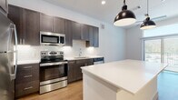 This single-floor unit is designed with a contemporary aesthetic, combining functionality and modern finishes throughout. The kitchen serves as a central highlight, featuring dark espresso-toned shaker cabinetry that extends to the ceiling, providing generous storage. A white subway tile backsplash contrasts cleanly against the cabinetry, while white quartz countertops extend across the perimeter and onto a sizable center island. A full suite of stainless steel appliances includes a refrigerator, electric range, over-the-range microwave, and dishwasher. Two matte black pendant lights hang above the island, adding a refined industrial touch. Light hardwood-style flooring runs throughout the main living areas, enhancing the bright, open feel of the space.

The bathroom continues the cohesive design language, showcasing matching dark cabinetry paired with a dual-sink vanity topped with white quartz. Framed mirrors with bar lighting provide ample illumination. A full-size soaking tub with white subway tile surround completes the space.

A dedicated laundry area includes a stacked washer and dryer unit alongside open built-in shelving, offering practical storage. The overall layout, visible in the 3D model, reflects an efficient, well-organized floor plan suited for comfortable everyday living.