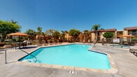 Located at 532 Broadway in El Cajon, California, this outdoor amenity area showcases a generously sized, irregularly shaped swimming pool with crystal-clear blue water, framed by a broad concrete pool deck. The deck is well-appointed with multiple chaise lounges and seating areas shaded by vibrant orange market umbrellas, providing ample space for relaxation and socializing under the Southern California sun. Adjacent to the main pool is a dedicated spa area featuring a bubbling hot tub, complemented by additional outdoor dining furniture and shading. The surrounding landscape is lush with mature palm trees, tropical plantings, and ornamental shrubs, enhancing the resort-like atmosphere. A wrought iron perimeter fence encloses the space, ensuring safety while maintaining open sightlines to the greenery beyond. The backdrop features warm-toned, earth-hued multi-story residential buildings with tile rooflines and balconies, adding architectural character to the setting. The overall ambiance is bright, inviting, and well-maintained, offering a comfortable outdoor retreat suited to the warm climate of the El Cajon area.