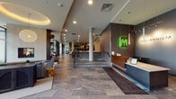 This contemporary lobby and amenity level presents a cohesive, upscale environment defined by clean lines, high-end materials, and thoughtful layout. The main entrance features floor-to-ceiling glass curtain walls with dark metal framing, flooding the entry vestibule with natural light and offering views of the surrounding streetscape. Large-format stone tile flooring in muted gray tones extends throughout the circulation corridors, anchoring the space with a sense of solidity and refinement.

At the heart of the lobby, a front desk is accented by a distinctive illuminated green neon art installation, creating a striking visual focal point. Concrete structural columns punctuate the open floor plan, lending an industrial undertone to the otherwise polished aesthetic. A split-level design connects the entry zone to an elevated corridor via wide stairs with glass-panel railings and dark metal handrails.

The lounge area features soaring ceilings, a floor-to-ceiling stone fireplace with an integrated flat-screen television, and oversized windows framing outdoor views. Comfortable seating arrangements and a sculptural globe pendant light contribute to an inviting atmosphere. Adjacent spaces include a glass-enclosed retail or market area with dark wood flooring, and an elevator bank finished in stainless steel with herringbone-patterned carpet flooring. A fitness facility is also accessible from the main corridor via glass-walled partitions.