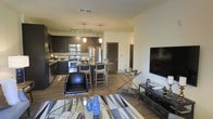 This single-floor residence showcases an open-concept design that integrates the living, dining, and kitchen areas into one cohesive and functional space. The kitchen is outfitted with dark espresso cabinetry, stainless steel appliances — including a refrigerator, microwave, and range — and a stylish tile backsplash. Pendant lighting hangs above a granite-topped kitchen island with bar seating, providing both a casual dining option and additional prep space. Track lighting overhead ensures the kitchen remains well-lit throughout. The living area features hardwood-style flooring that extends throughout the open space, grounding the layout with warmth and continuity. A large flat-screen television is mounted opposite a seating arrangement that includes a leather accent chair and a glass-topped coffee table, styled with decorative accessories. A geometric area rug defines the living zone, adding texture and visual interest. Crown molding along the ceiling provides a refined architectural detail. The overall ambiance is modern and well-appointed, with a neutral palette complemented by bold, dark cabinetry and contemporary furnishings that create a polished, move-in-ready feel.