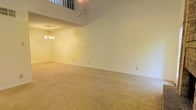 This two-story space makes a strong architectural statement with its soaring double-height ceiling in the main living area, creating an expansive and airy atmosphere. A white-railed upper-level overlook peers down into the living space below, adding a sense of openness and vertical drama that defines the character of the home. The living area is generously proportioned, finished with wall-to-wall plush carpeting in a warm neutral tone that adds comfort underfoot and contributes to the overall inviting ambiance. A classic multi-arm chandelier serves as a central decorative focal point, suspended beneath the tall ceiling and providing ambient illumination to the space. The walls are finished in a clean, light neutral tone, offering a versatile backdrop adaptable to a variety of interior styles. White baseboard trim runs throughout, providing a crisp, polished contrast to the carpeted floors. The two-story layout adds both functional living space and architectural interest, with the upper level contributing to the sense of scale when viewed from the main floor. Overall, the space conveys a comfortable, traditional aesthetic with timeless design elements suited for everyday living.