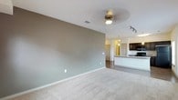 This single-floor space offers a clean, open-concept layout that seamlessly connects the living area to the kitchen. The living area is finished with plush, neutral-toned carpeting and painted in a soft sage green, creating a calm and welcoming ambiance. A ceiling fan with an integrated light fixture is centrally positioned overhead, providing both comfort and illumination. The transition into the kitchen and dining area is marked by a shift to hard flooring, adding a practical contrast to the soft living space. The kitchen is well-appointed with dark espresso cabinetry, a built-in microwave, and a countertop that extends into a breakfast bar — ideal for casual dining or entertaining. Track lighting above the kitchen adds focused, functional brightness to the workspace. A pendant light above the bar area adds a refined touch. The layout flows naturally toward a hallway that leads to additional rooms, suggesting a thoughtful floor plan designed for everyday living. White trim and neutral wall tones throughout lend the space a bright, cohesive aesthetic that is both modern and versatile.