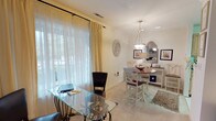 This single-floor space showcases an open and versatile layout, combining a functional home office area with an adjacent dining space. The office area is anchored by a glass-topped desk with an ornate metal base and a dark upholstered chair, positioned near a large window dressed with sheer white and cream drapery panels that allow abundant natural light to filter through. Views of greenery outside enhance the bright, airy feel of the room.

The dining area flows naturally from the office space and features a round wooden table paired with elegant white-painted chairs with warm wood accents, set atop a patterned area rug over neutral carpeted flooring. A classic pendant light with a bell-shaped shade hangs above the dining table, providing warm, focused illumination. Against the wall, a white console table with decorative mirrors and framed botanical artwork adds a refined, traditional character to the space.

The walls are finished in a soft, warm white tone, and recessed lighting complements the pendant fixture overhead. The overall ambiance is light, welcoming, and thoughtfully arranged, blending practicality with classic elegance throughout the combined living areas.