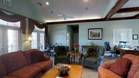 This single-floor space exudes a warm, classic ambiance anchored by a generously sized living area designed for both comfort and sophistication. The room features rich, dark wood beam accents that frame the ceiling, adding architectural character and depth. A ceiling fan sits centrally overhead, complementing the recessed lighting that illuminates the space evenly throughout. Large windows dressed with ornate dark drapery allow natural light to pour in, creating a bright and inviting atmosphere. The seating arrangement is thoughtfully composed, featuring plush tufted armchairs with nailhead trim, a deep-cushioned sofa in warm earth tones, and a solid wood coffee table as the centerpiece. A decorative bowl and reading materials rest on the table, adding a lived-in, welcoming feel. In the background, a compact dining or gathering area with wrought iron furniture is visible, along with French doors that offer a connection to the exterior. A framed landscape painting adorns one wall, contributing to the refined, traditional aesthetic. A small desk or console near the far wall provides a functional workspace within the room. The overall design blends comfort and elegance, making this a versatile and inviting space suited for relaxation or casual gatherings.