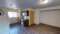 This single-floor unit offers a practical and efficient layout designed to maximize its compact footprint. The open-concept living and kitchen area creates a sense of spaciousness, with dark hardwood-style laminate flooring running throughout and providing a warm, cohesive aesthetic.

The kitchen is positioned along one wall and features dark espresso-toned cabinetry offering ample storage, complemented by granite-look countertops. White appliances, including an electric range and over-the-range microwave, are neatly integrated into the cabinetry. A window above the kitchen area allows natural light to filter into the space.

The living area is open and unobstructed, benefiting from flush-mount ceiling lighting and neutral wall tones that keep the atmosphere bright and versatile. Warm wood-toned baseboards add a finishing detail that ties together the flooring and walls. A dedicated interior door leads to additional areas of the unit, and a floor vent indicates a forced-air climate system. The overall design is clean and functional, well-suited for everyday living.