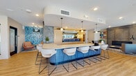 This thoughtfully designed single-floor communal space offers a variety of distinct zones suited for socializing, relaxing, and focused gatherings. The centerpiece is a generously sized bar-height island clad in vibrant cobalt blue tile with a clean white countertop, surrounded by contemporary white upholstered barstools with sleek metal legs. Globe pendant lights hang above, complementing the warm light wood flooring that flows throughout the entire space.

Adjacent to the island, a lounge area features plush gray sofas, a round coffee table, and a wall-mounted flat-screen TV framed in light wood paneling. A textured herringbone accent wall adds visual depth, while sliding glass-panel doors provide access to a private room. Nearby, a cozy fireplace nook showcases a dramatic floor-to-ceiling concrete surround with a linear gas fireplace, flanked by open wood shelving and comfortable seating.

A dedicated kitchenette area offers warm wood cabinetry, a utility sink, and open shelving for additional functionality. A bold tropical-print wallpapered accent wall introduces color and energy to the social zone. Large black-framed glass doors at one end of the space flood the interior with natural light and open directly onto a spacious outdoor patio, seamlessly connecting the interior and exterior environments.
