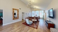 Located at 19 Landmark Dr in Columbia, SC, this thoughtfully designed single-floor community space offers a generous layout encompassing a living room, combined dining room and kitchen, game room, office, and hallway — all unified by warm hardwood-style flooring and a neutral, inviting color palette.

The living room is a standout area, boasting soaring vaulted ceilings and dramatic floor-to-ceiling windows that flood the space with natural light and offer views of the surrounding greenery. Elegant candelabra chandeliers add a refined ambiance, complemented by a large decorative mirror that enhances the sense of openness.

The combined dining room and kitchen is equally impressive. The kitchen features dark cabinetry, stainless steel appliances, granite countertops, a striking mosaic tile backsplash, and a curved island with bar seating beneath pendant lighting. The adjacent dining area is bathed in natural light from large windows overlooking a landscaped exterior, and offers multiple seating arrangements including a formal dining table and high-top tables.

The game room provides a recreational retreat, centered around a full-size pool table and accented with playful wall décor. The office is lined with bookshelves and features large windows. A welcoming entry hallway with sheer-curtained windows completes this versatile and stylishly finished space.