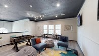 This single-floor community space is thoughtfully designed for both recreation and relaxation, offering a versatile layout that accommodates a variety of activities. The room is immediately distinguished by its bold, dark textured ceiling with recessed and track lighting, creating a modern contrast against the crisp white wainscoted walls and light wood-style flooring throughout.

The central lounge area features comfortable blue upholstered accent chairs and bean bag seating arranged atop a vibrant abstract area rug in teal, yellow, and grey tones. A full-size foosball table anchors the gaming zone, providing an engaging focal point for social interaction. Two wall-mounted flat-screen TVs are positioned for easy viewing from the lounge seating.

Along one wall, a dedicated computer workstation area is outfitted with desks and bright yellow chairs, offering a practical space for productivity. A leather bench with decorative pillows provides additional casual seating nearby.

A separate dining nook lines the exterior wall, featuring rows of windows that allow ample natural light to fill the space and offer views of the surrounding greenery. Bistro-style tables paired with metal industrial chairs create a café-like atmosphere ideal for casual gatherings or individual use. Framed artwork decorates the walls, adding character to the overall design. A ceiling fan ensures year-round comfort throughout the space.