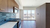 Located at 2215 NW Quimby St in Portland, Oregon, this single-floor unit offers a thoughtfully designed open layout that integrates the kitchen and living areas seamlessly. Rich hardwood-style flooring runs throughout, providing a warm contrast to the clean, neutral wall tones and modern finishes found across the space.

The kitchen is a standout feature, equipped with stainless steel appliances including a gas range, built-in microwave, and refrigerator. Deep walnut-toned upper cabinetry pairs with light countertops and a striking blue tile backsplash, adding a bold design element. A floor-to-ceiling picture window anchors the living area, flooding the space with natural light and framing expansive views of the Portland skyline and surrounding cityscape.

The bedroom area is well-proportioned and includes a large sliding panel partition, offering a flexible and modern approach to room division. The bathroom features a full soaking tub and shower combination with crisp white tile surround, accented by warm brass fixtures and a marble-style vanity countertop, creating a clean, contemporary aesthetic.

The entry corridor provides a practical transition into the living space, with additional storage integrated along the hallway. Overall, the unit delivers a cohesive, modern urban living experience with quality finishes and notable natural light.
