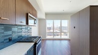 Located at 2215 NW Quimby St in Portland, Oregon, this single-floor unit offers a thoughtfully designed open layout that integrates the kitchen and living areas seamlessly. Rich hardwood-style flooring runs throughout, providing a warm contrast to the clean, neutral wall tones and modern finishes found across the space.

The kitchen is a standout feature, equipped with stainless steel appliances including a gas range, built-in microwave, and refrigerator. Deep walnut-toned upper cabinetry pairs with light countertops and a striking blue tile backsplash, adding a bold design element. A floor-to-ceiling picture window anchors the living area, flooding the space with natural light and framing expansive views of the Portland skyline and surrounding cityscape.

The bedroom area is well-proportioned and includes a large sliding panel partition, offering a flexible and modern approach to room division. The bathroom features a full soaking tub and shower combination with crisp white tile surround, accented by warm brass fixtures and a marble-style vanity countertop, creating a clean, contemporary aesthetic.

The entry corridor provides a practical transition into the living space, with additional storage integrated along the hallway. Overall, the unit delivers a cohesive, modern urban living experience with quality finishes and notable natural light.