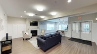 This single-floor community space is thoughtfully designed to foster comfort and social connection, blending modern aesthetics with a warm, welcoming ambiance throughout. The main lounge area serves as the heart of the space, featuring a classic white fireplace mantel with a wall-mounted flat-screen TV, elegant wainscoting panels, and a seating arrangement of plush navy sofas and upholstered armchairs arranged over a textured area rug. Light wood-tone flooring flows seamlessly throughout, complementing the neutral warm-gray and white color palette. A secondary lounge nook mirrors this inviting atmosphere with additional armchairs and a cozy fireplace, ideal for quieter gatherings. The open-plan layout extends into a bar-style counter area highlighted by a striking dark subway tile accent wall, pendant lighting, and large windows that invite natural light and outdoor views. The fully equipped kitchen features rich wood cabinetry, stainless steel appliances including a refrigerator, range, microwave, and dishwasher, paired with a coordinating tile backsplash. Adjacent to the kitchen, a spacious dining area offers multiple tables with cross-back chairs, well-lit by overhead fixtures, and accented with framed artwork. The space is practical, polished, and well-suited for communal living and entertaining.